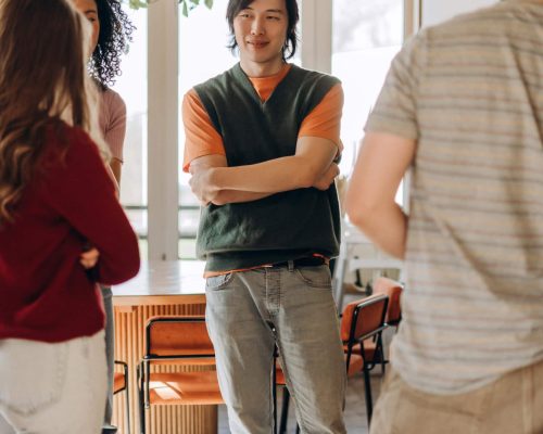 Young Japanese man leading discussion with creative team in modern office Young Japanese man with crossed arms stands confidently amidst colleagues in a brightly lit, modern office space, engaging in collaborative discussion and idea exchange
